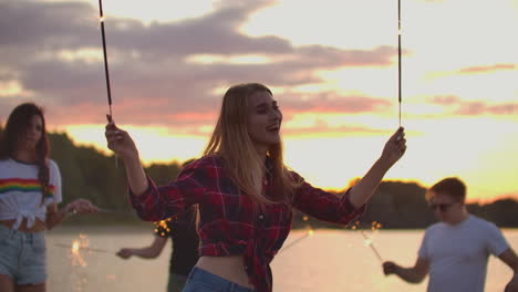 The-blonde-woman-in-red-plaid-shirt-and-denim-shorts-is-dancing-with-big-bengal-lights-in-her-hands-on-the-beach-with-her-friends.-This-is-beautiful-summer-evening-on-the-lake-coast-near-the-forest.